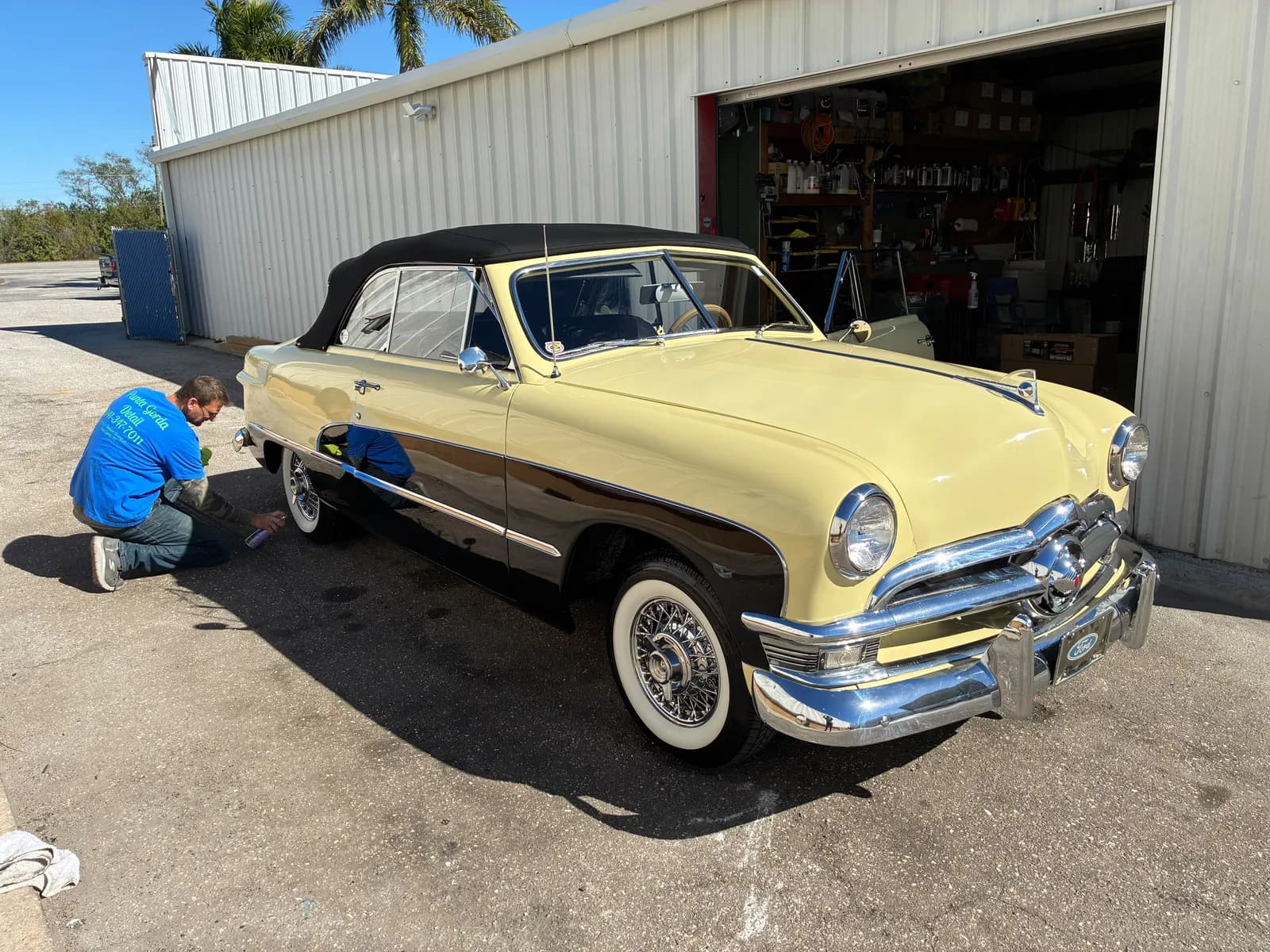 Detailer dressing the whitewall tires of a yellow and black 1950 Ford convertible outside the shop.